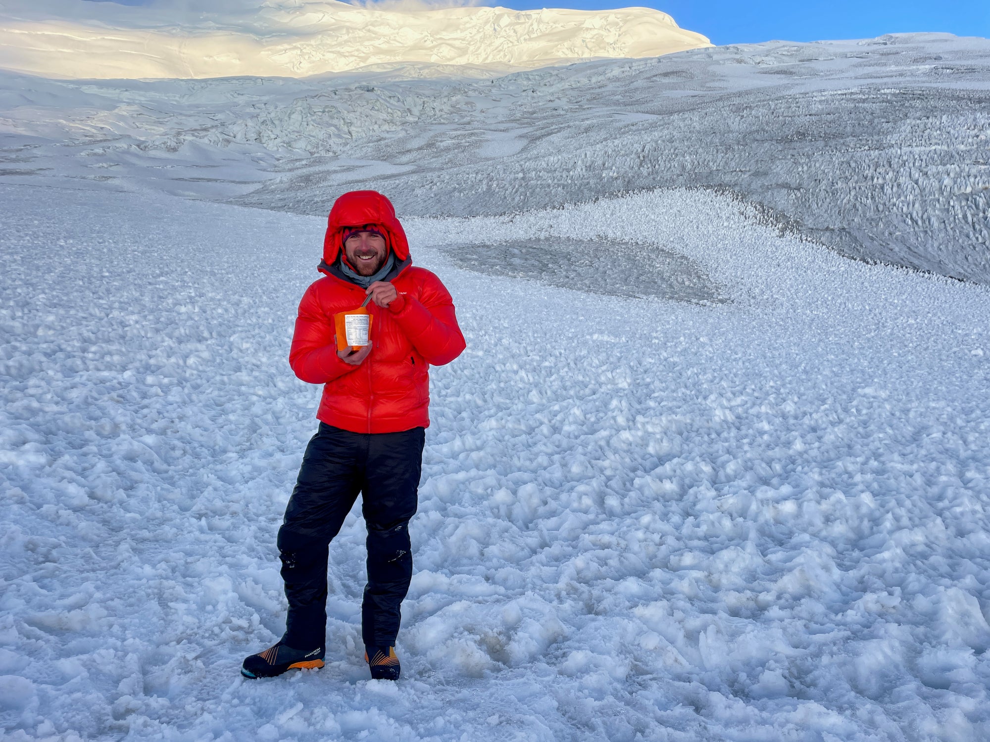 A man standing in a snow field wearing a bright red jacket eating an Expedition Foods freeze-dried meal
